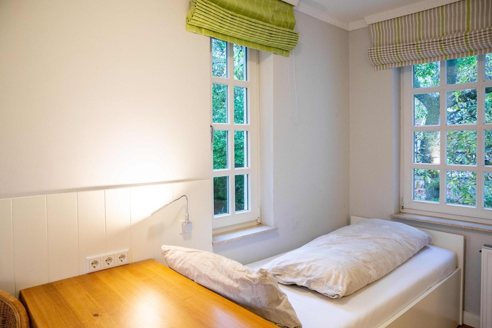 Schlafzimmer mit Bett, Schreibtisch und Fenstern mit Blick auf Haus und Grün.