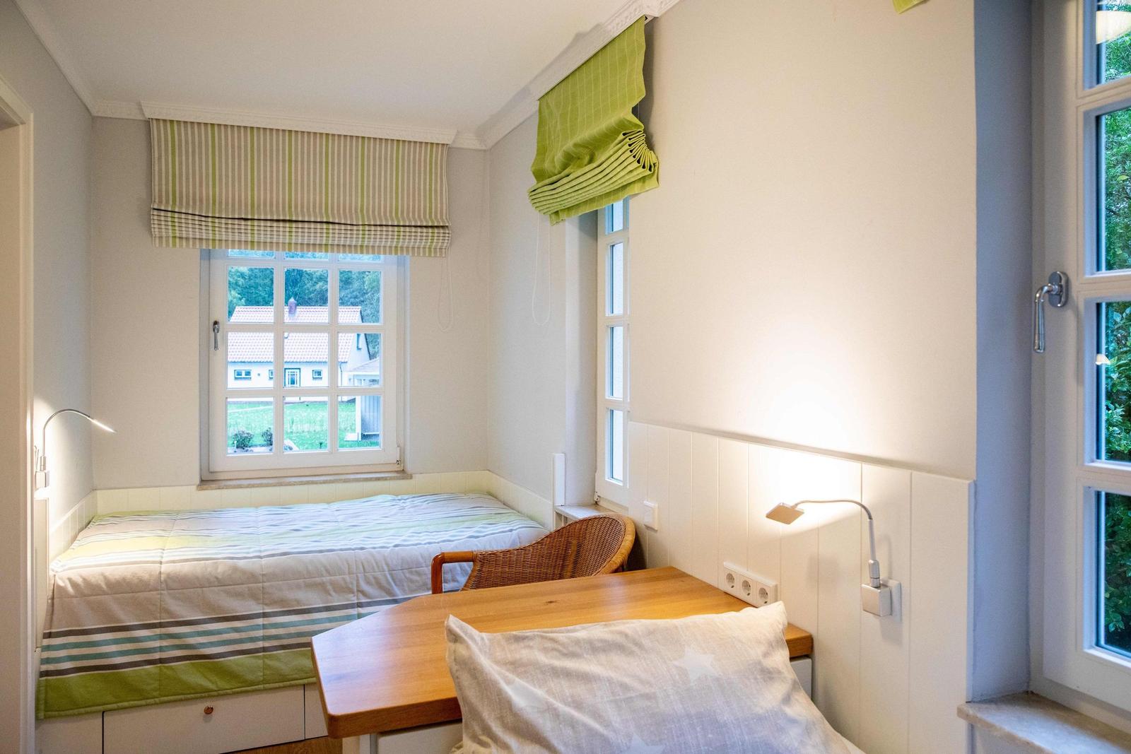Zimmer mit zwei Fenstern, Bett, Tisch und Stuhl. Grüne Rollläden und Holzmöbel.