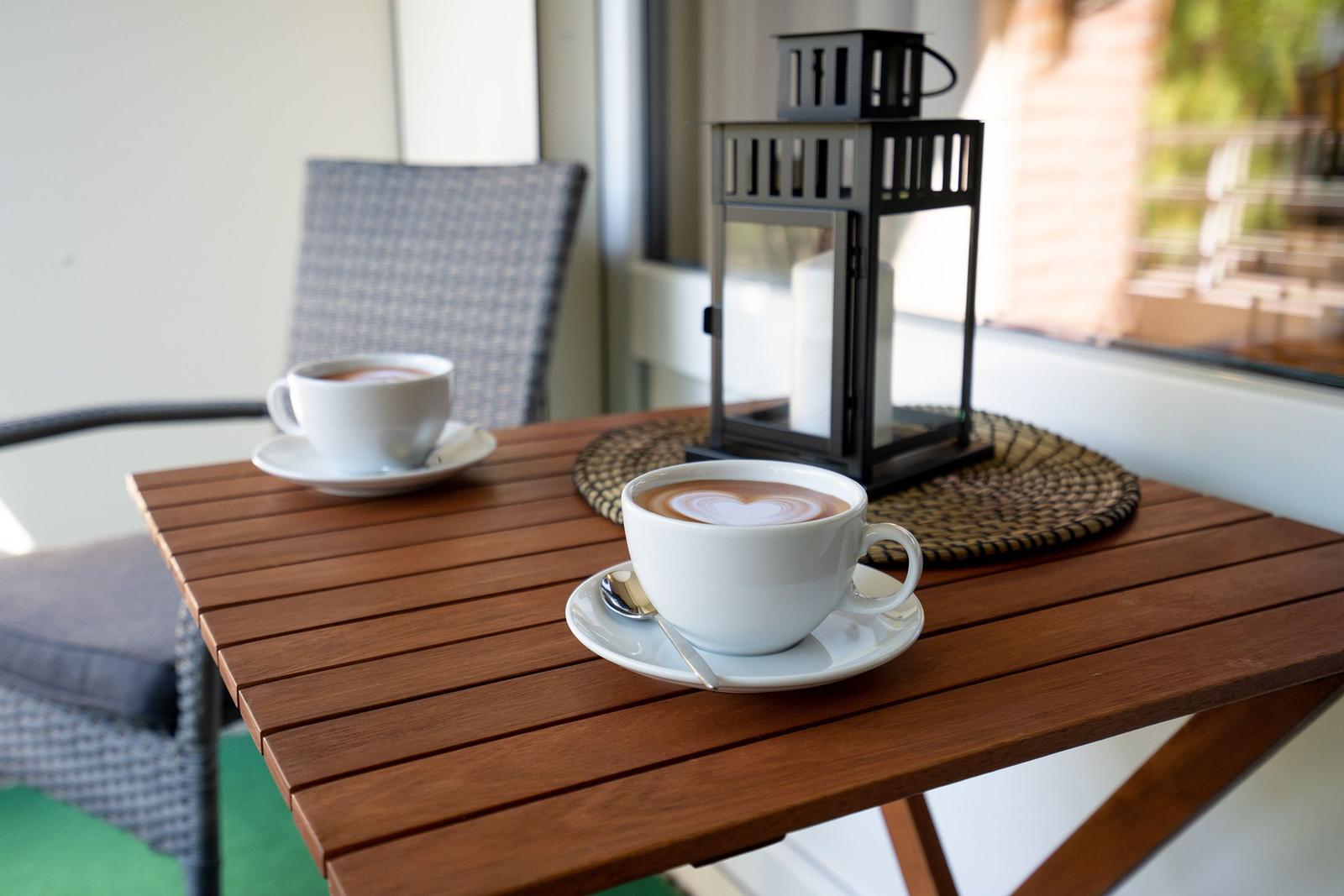 Terrasse mit grünem Sonnenschirm, zwei Stühlen und Tisch. Grünflächen und Gebäude im Hintergrund.