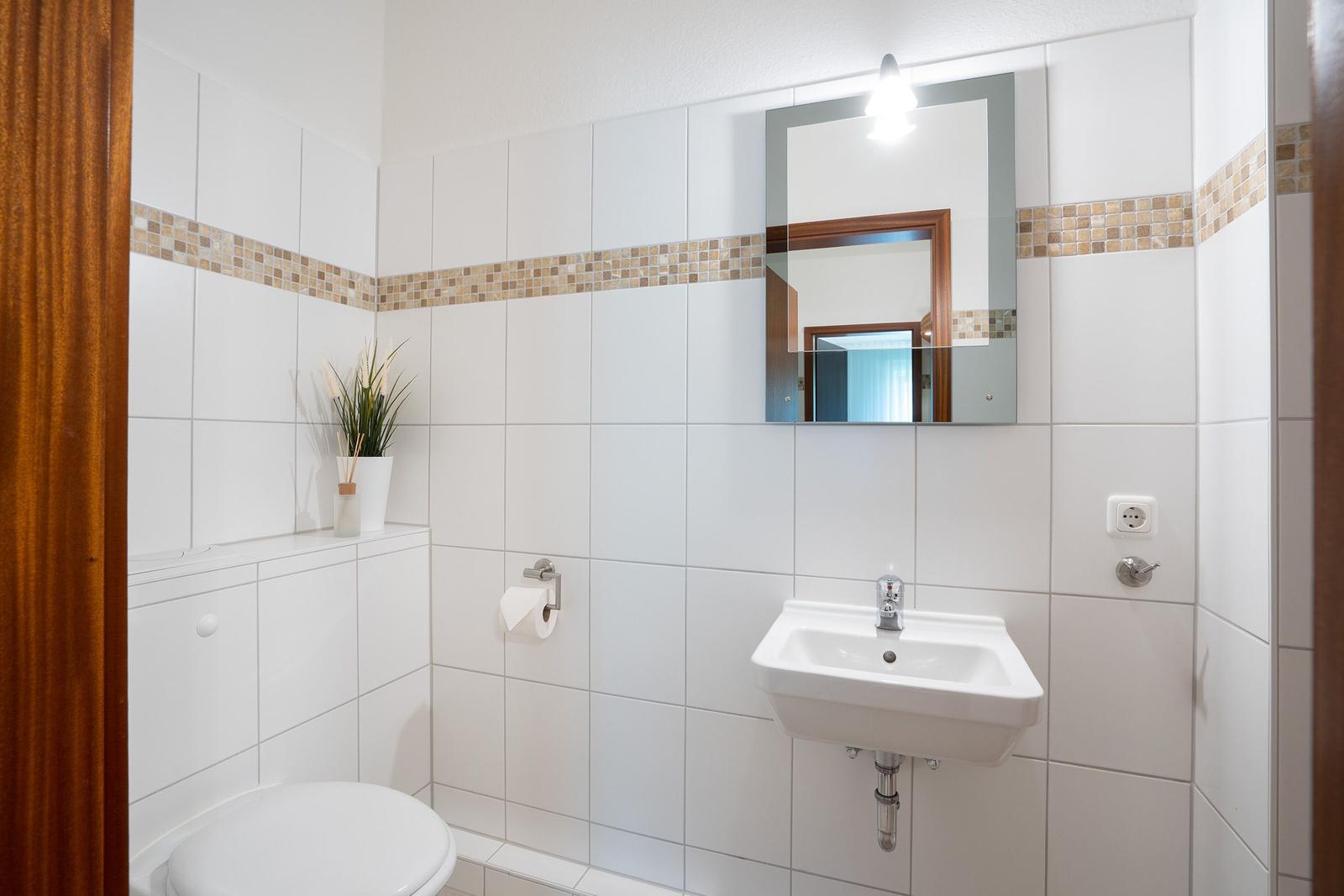 Badezimmer mit weißen Fliesen und Mosaikband. Pflanze und Aromalampe auf der Arbeitsfläche.