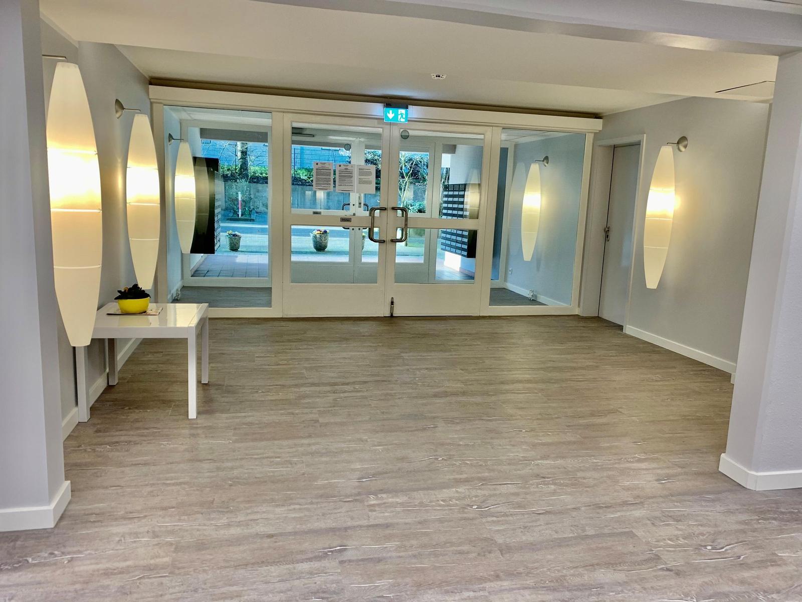 Helle Lobby mit Holzboden, Wandlampen und Tisch mit Pflanze. Lift im Hintergrund.
