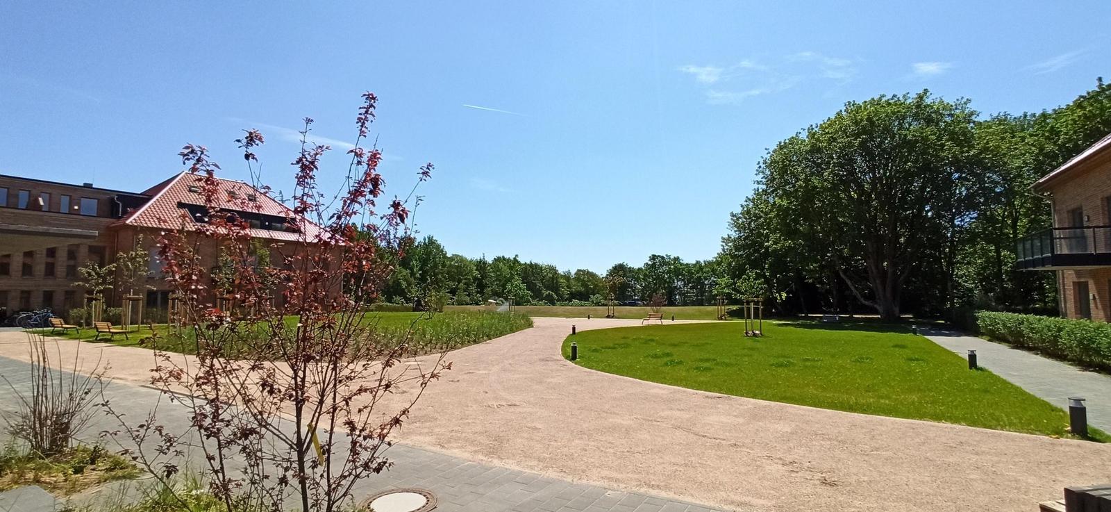 Zwei Gebäude mit roten Dächern und Balkonen umgeben einen gepflasterten Platz mit Grünfläche.