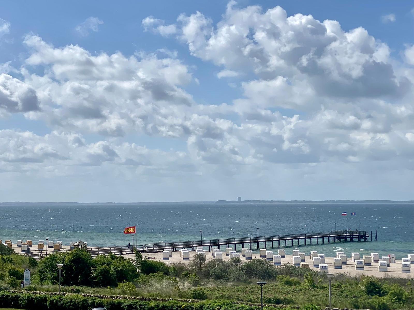 Meer mit Strand, Strandkörben und grüner Vegetation unter blauem Himmel mit Wolken.