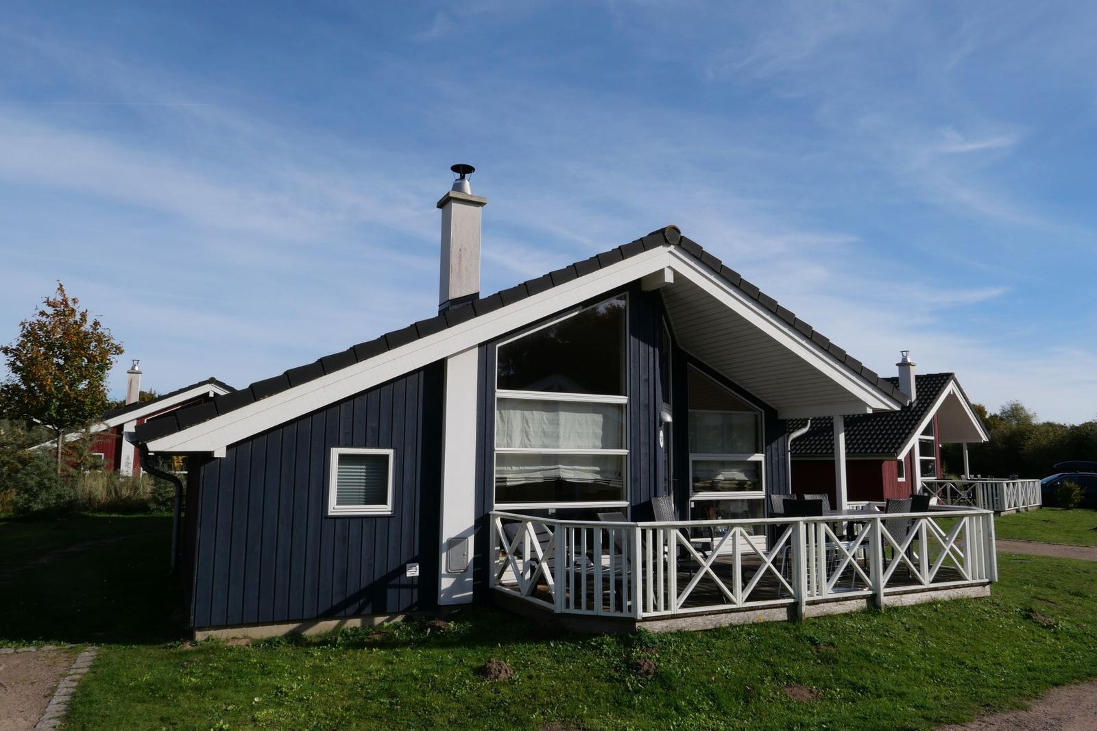 Blau gestrichenes Ferienhaus mit weißen Fensterrahmen und Terrasse. Hintergrund: weitere Häuser und Grünflächen.