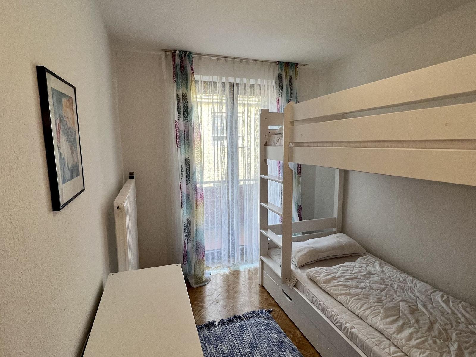 Zimmer mit Doppelstockbett, Stuhl und Schreibtisch. Tür führt zu Balkon.