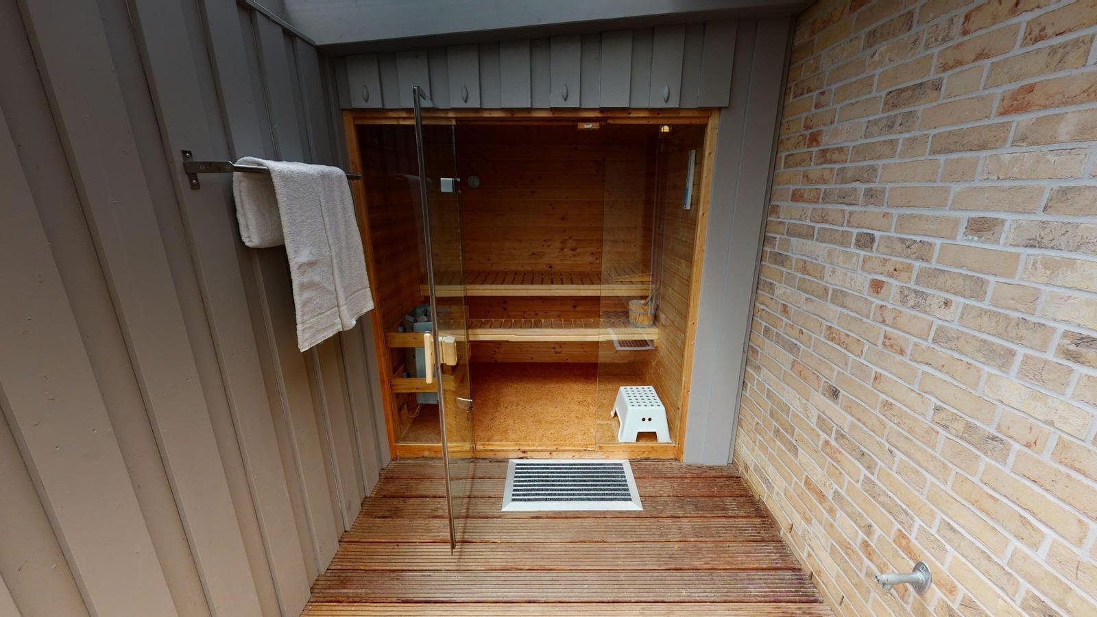 Sauna mit Holzbänken, Holzregal und Eimer mit Besen. Fenster zeigt Außenansicht.