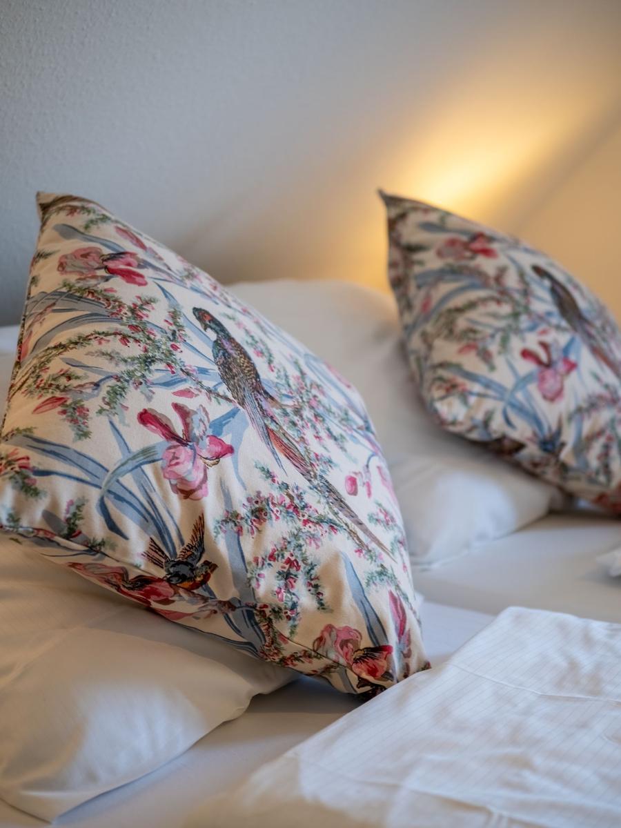 Schlafzimmer mit Bett, Schreibtisch, Stuhl und Wandbild. Fenster mit floralen Vorhängen.