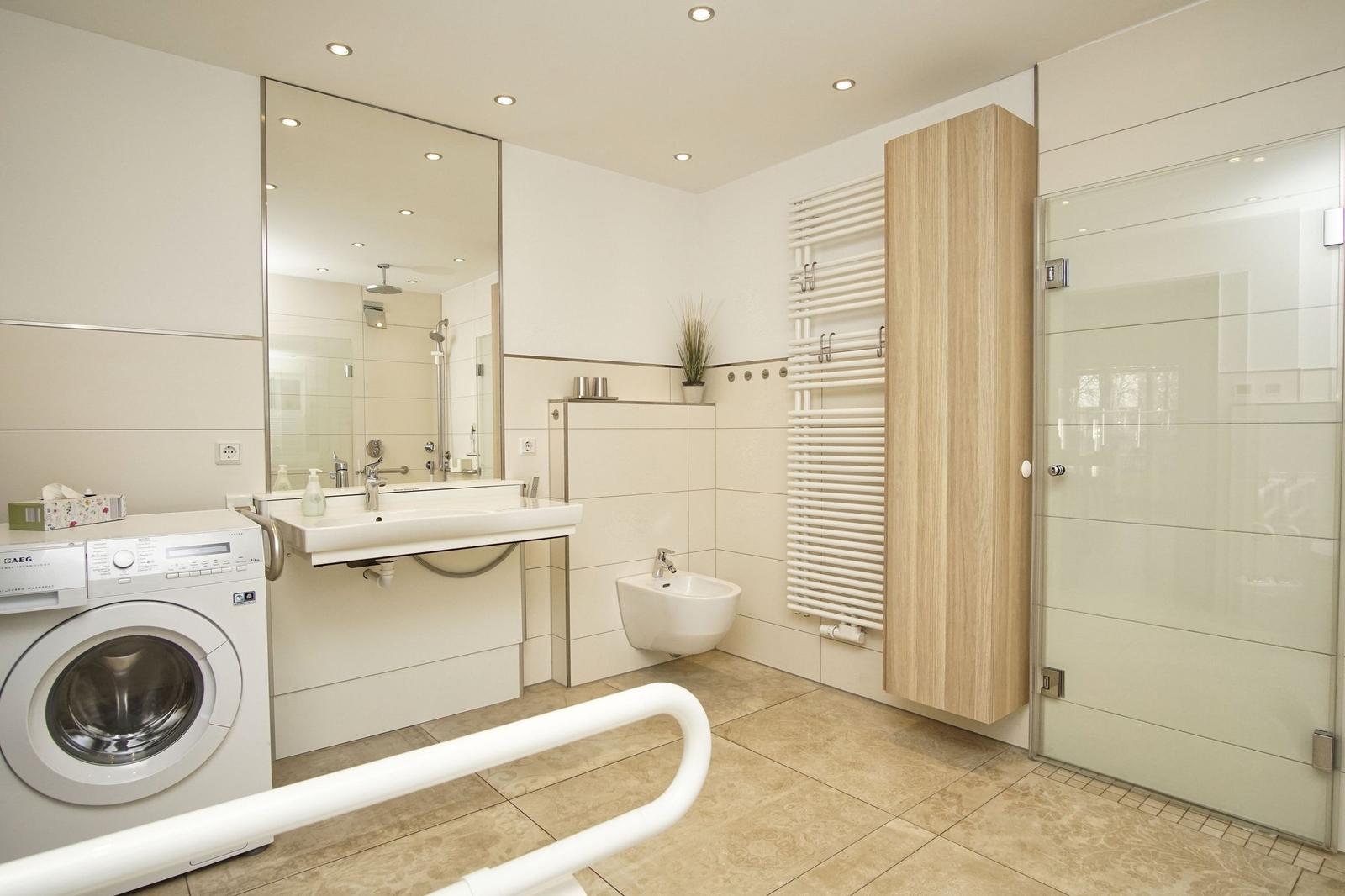 Badezimmer mit Dusche, WC und Waschbecken. Fliesenboden und Handlauf für barrierefreien Zugang.