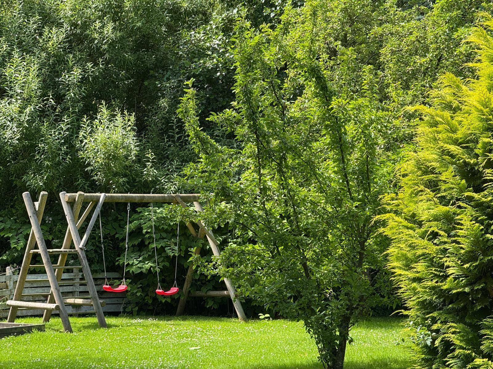 Holztisch mit Bänken, blauer Sonnenschirm und Hängemöbel im Garten.