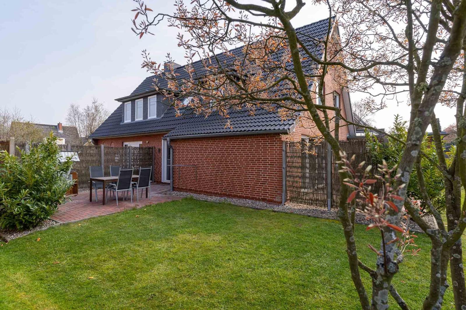 Terrasse mit Holztisch, Stühlen und Strandkorb. Hintergrund: Haus mit Ziegelfassade und Dach.