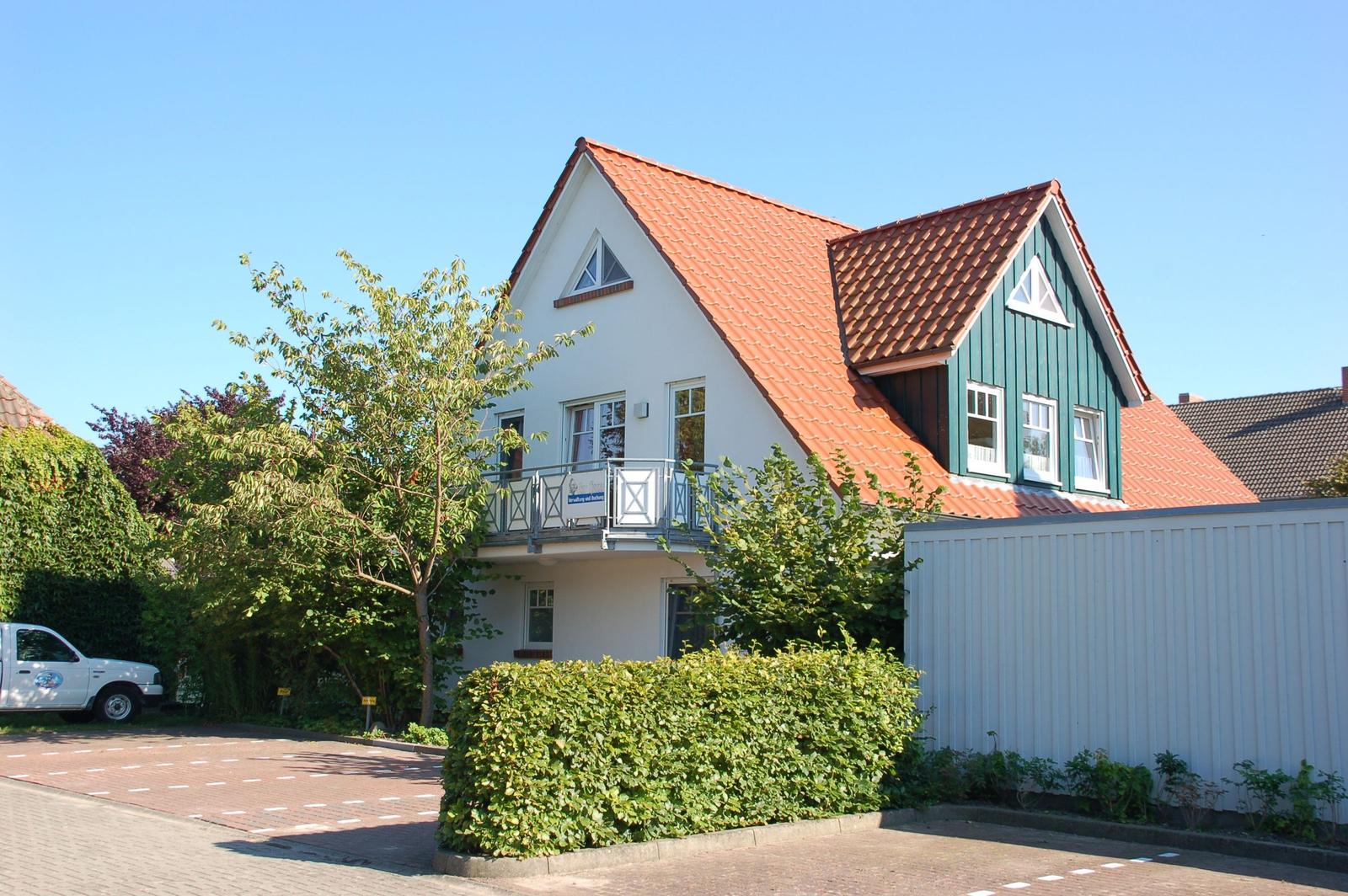 Wohnhaus mit Balkon, Gartenbereich und Liegestühlen. Holztor und grüne Bäume umgeben das Grundstück.