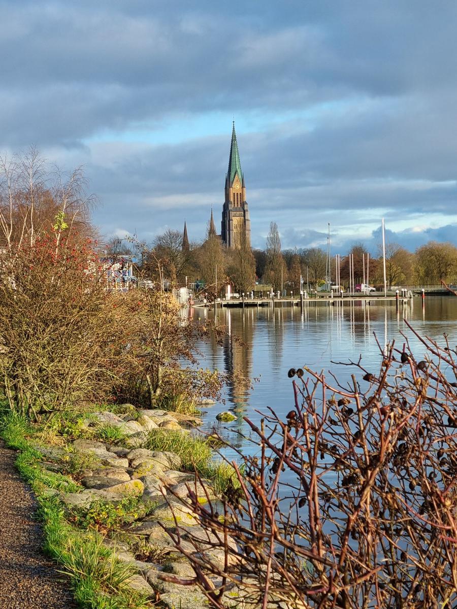 Bronzefigur an einem Flussufer mit Blick auf eine Kirche und Bootshäuser.