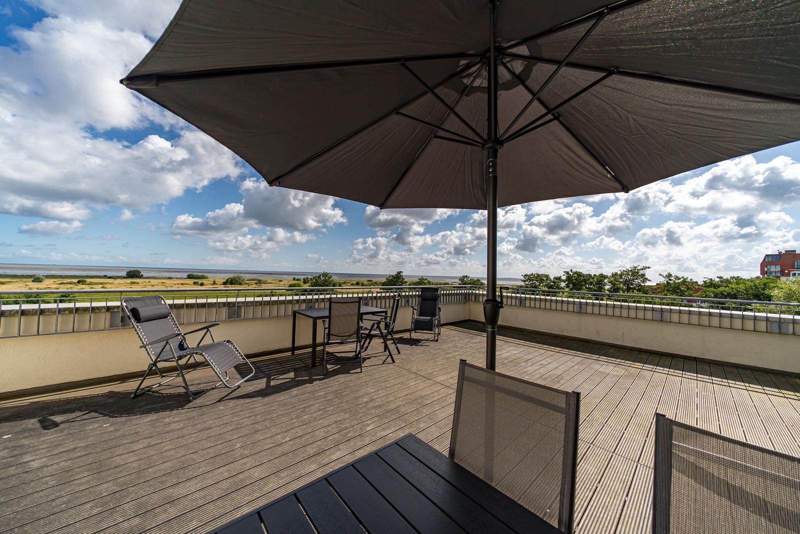 Terrasse mit Tischen, Stühlen und Sonnenschirm. Blick auf Stadt und Bäume unter blauem Himmel.