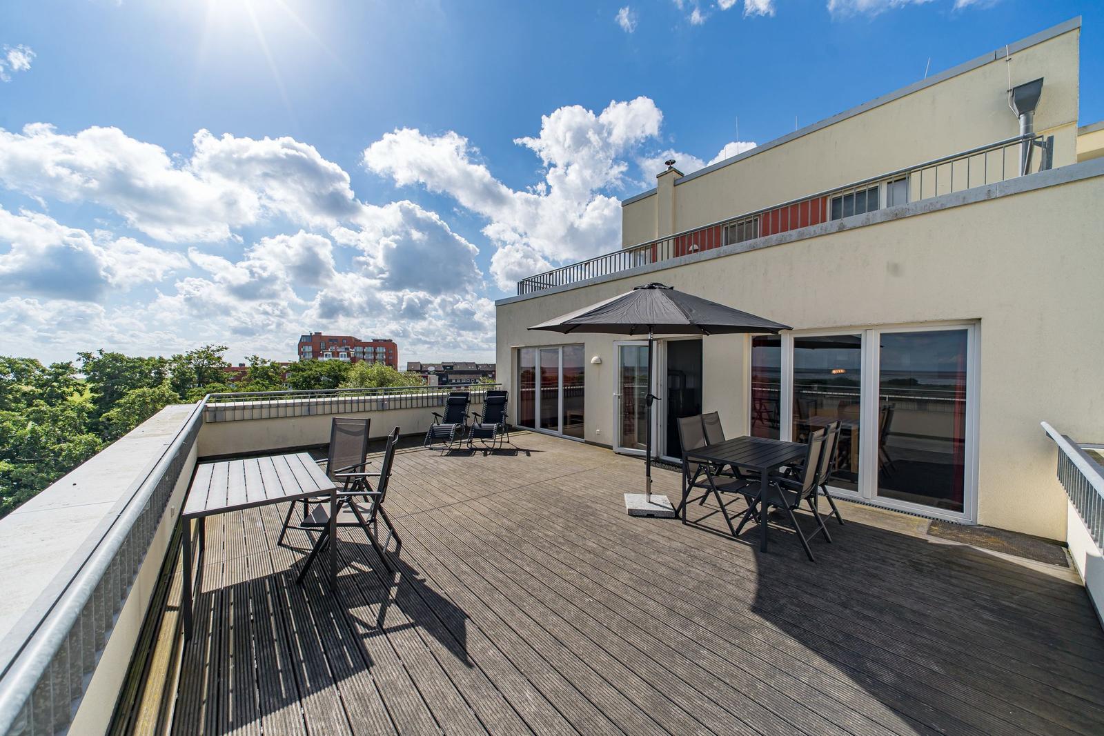 Terrasse mit Tisch, Stühlen und Sonnenschirm. Blick auf Felder und Wasser unter blauem Himmel mit weißen Wolken.