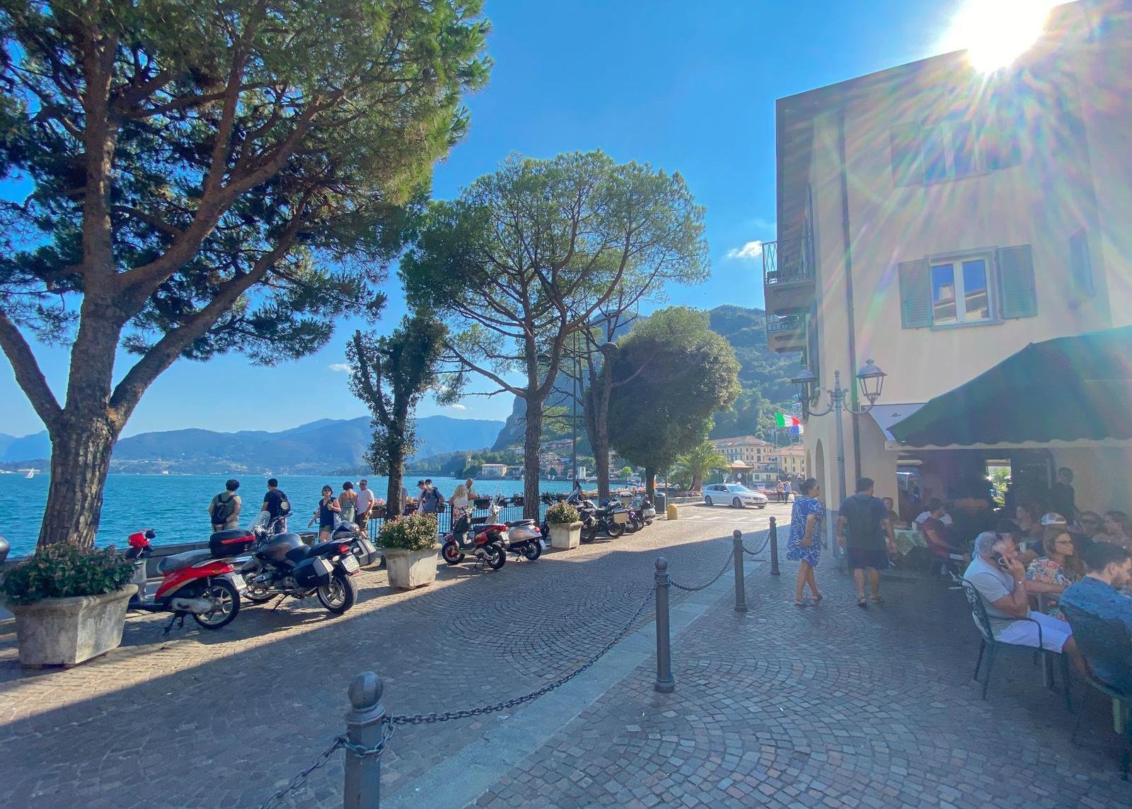 Pavement area near lake with trees, people walking, and café seating under umbrellas.