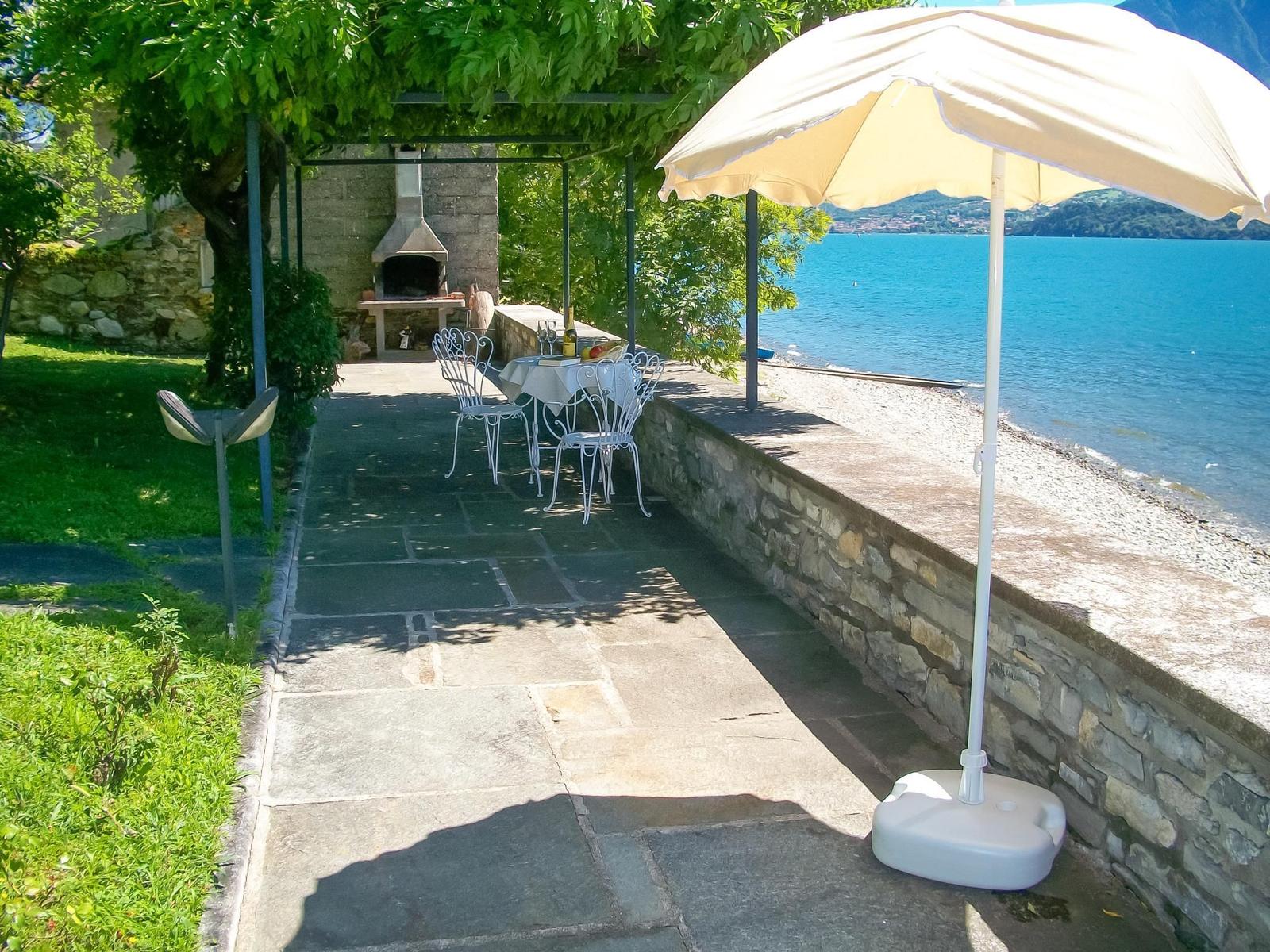 Garten mit Tisch und Stühlen unter Bäumen. Blick auf See mit Liegestühlen.