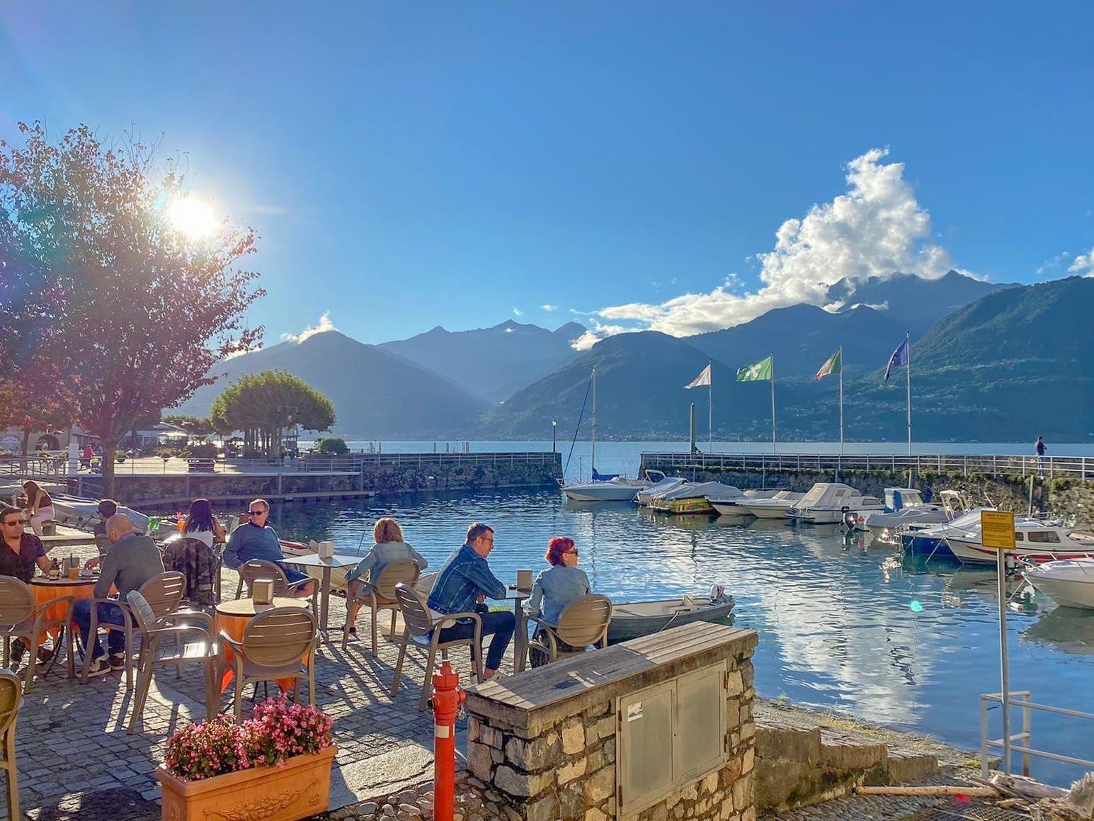 Sonnenlicht über gepflastertem Platz mit Bäumen, Sitzgelegenheiten und Blick auf See und Berge.