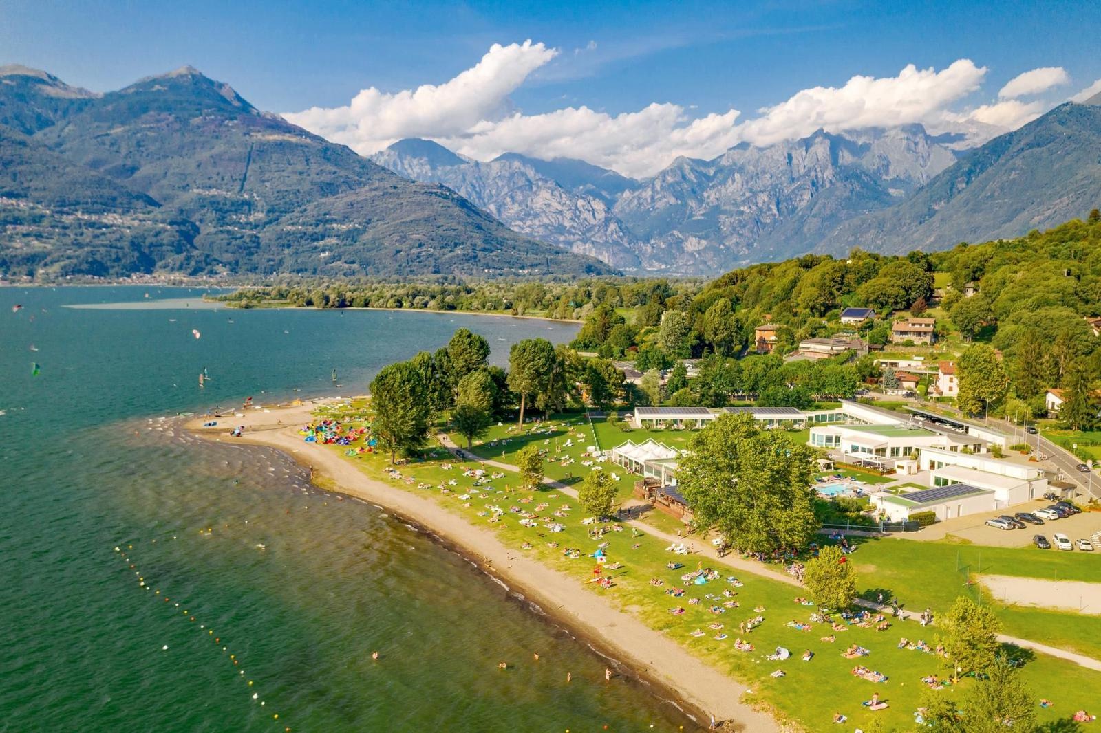 Grünes Ufer mit Menschen, Bergen und einem Segelflieger am See.
