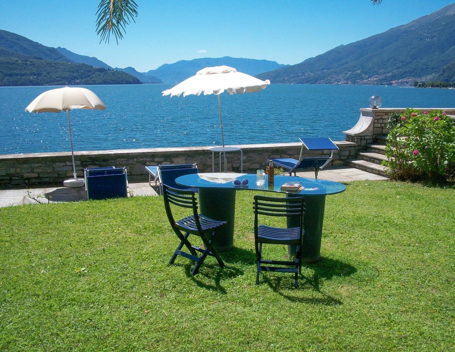 Terrasse mit Tisch, Stühlen und Sonnenschirm am See. Steinmauer und Bergblick.