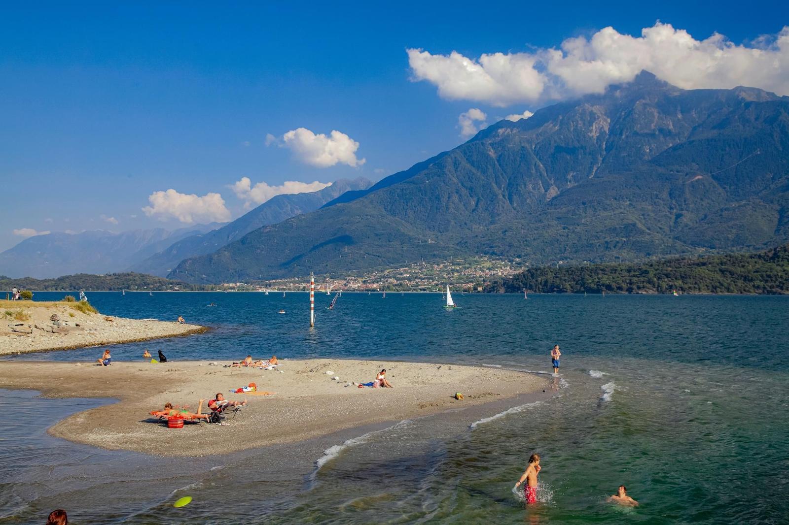 Grüner Rasen am See mit Liegestühlen unter einem Baum. Berge im Hintergrund.