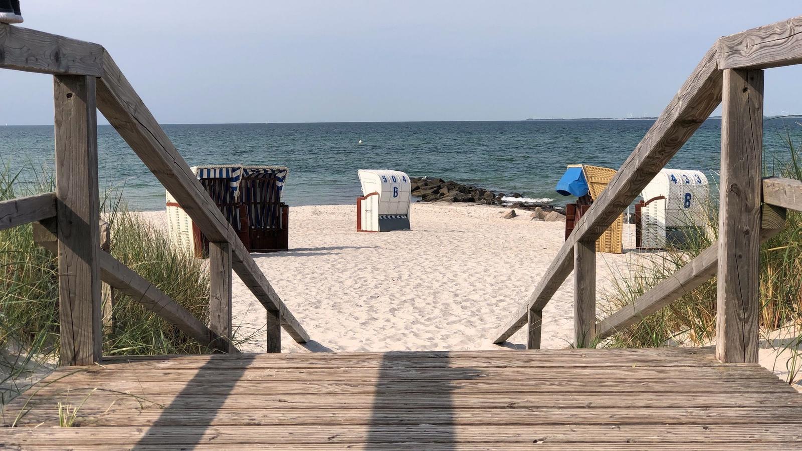 Strand mit Strandkörben, einem Gebäude und einer Person am Wasser.