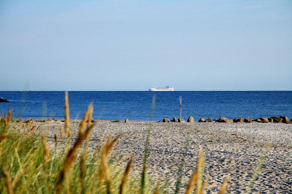 Drei Personen fahren mit Fahrrädern entlang eines Strandufers mit Blick auf einen Steg ins Meer.