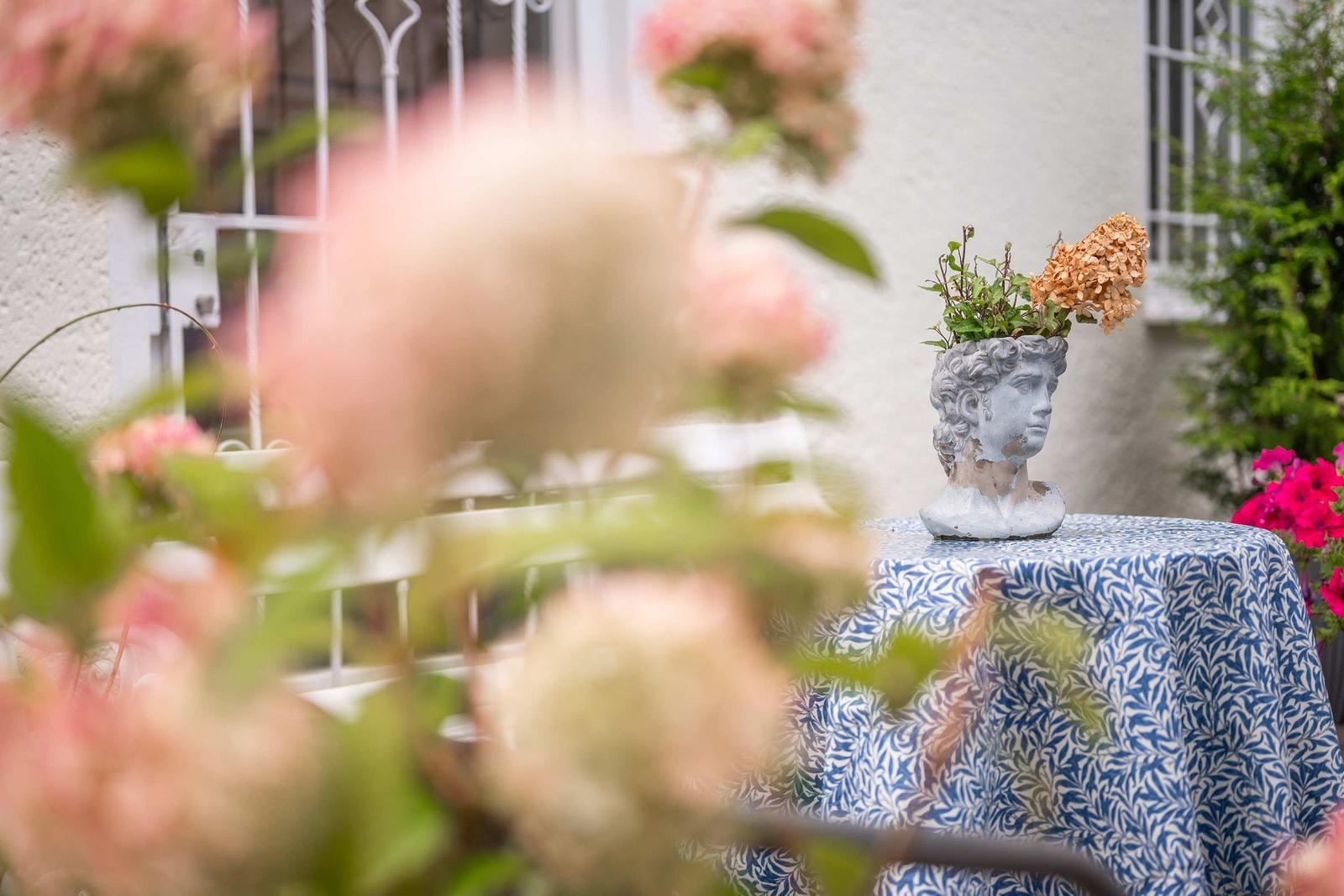 Backyard with paved ground, flowers, and tables. White house wall and balcony.