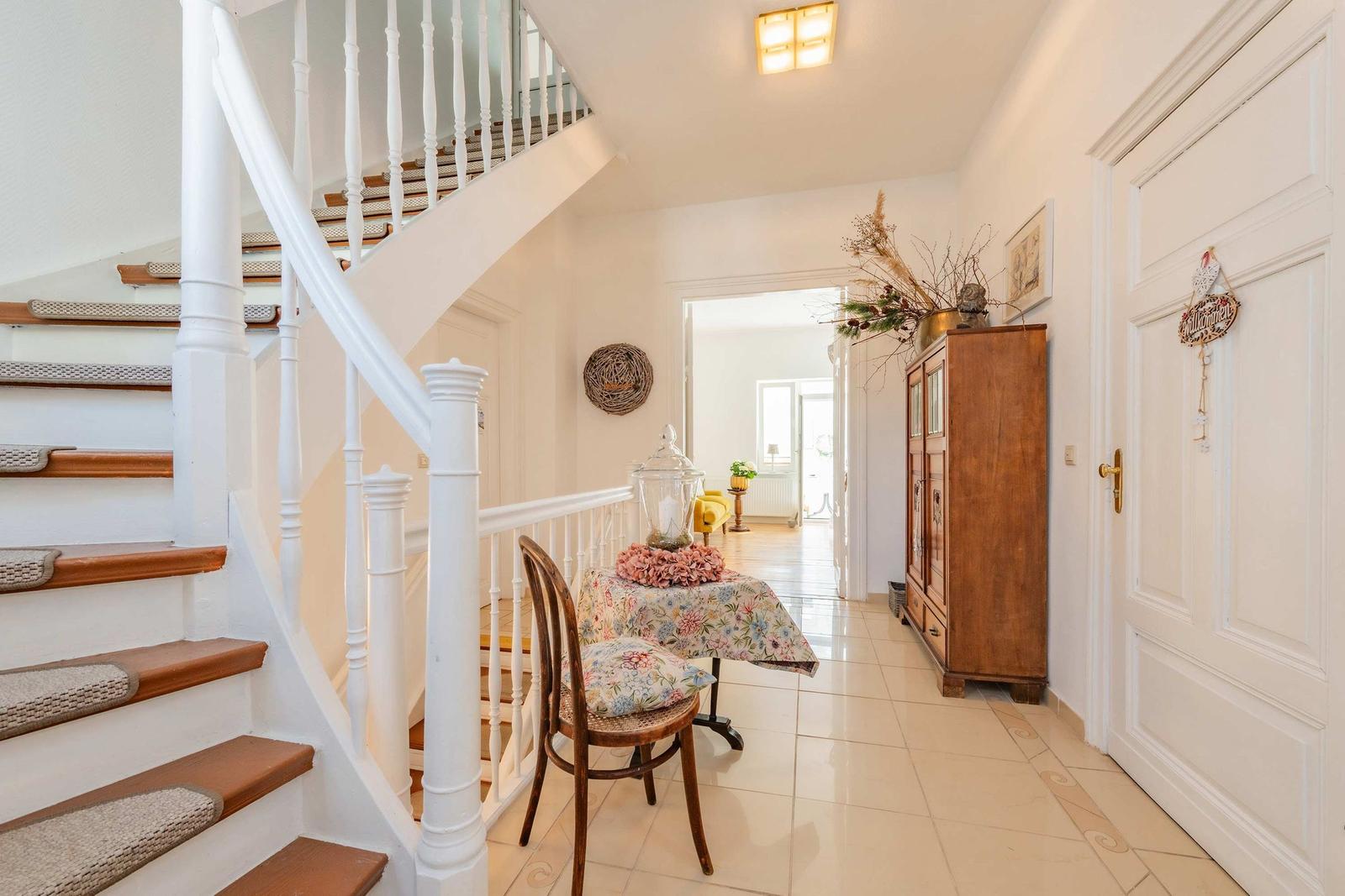 Entrance area with wooden chair, table with floral cloth, view into living room.