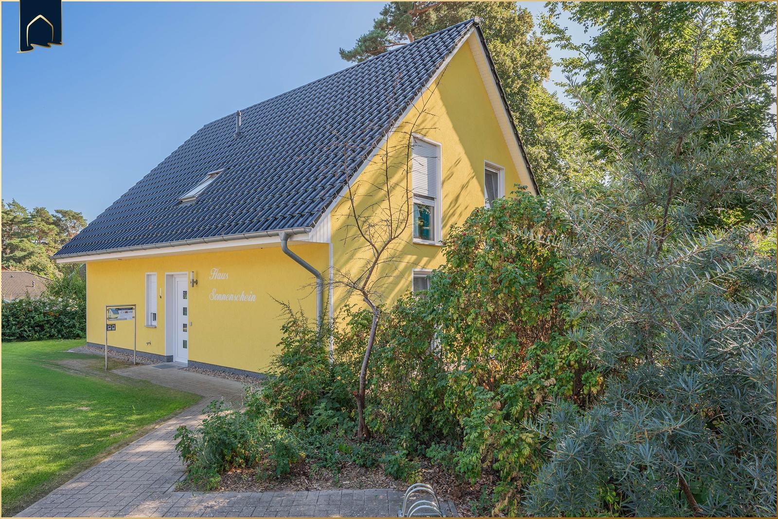 Gelbes Haus mit grauem Dach, Fenstern und Tür. Umgeben von Gras und Bäumen.
