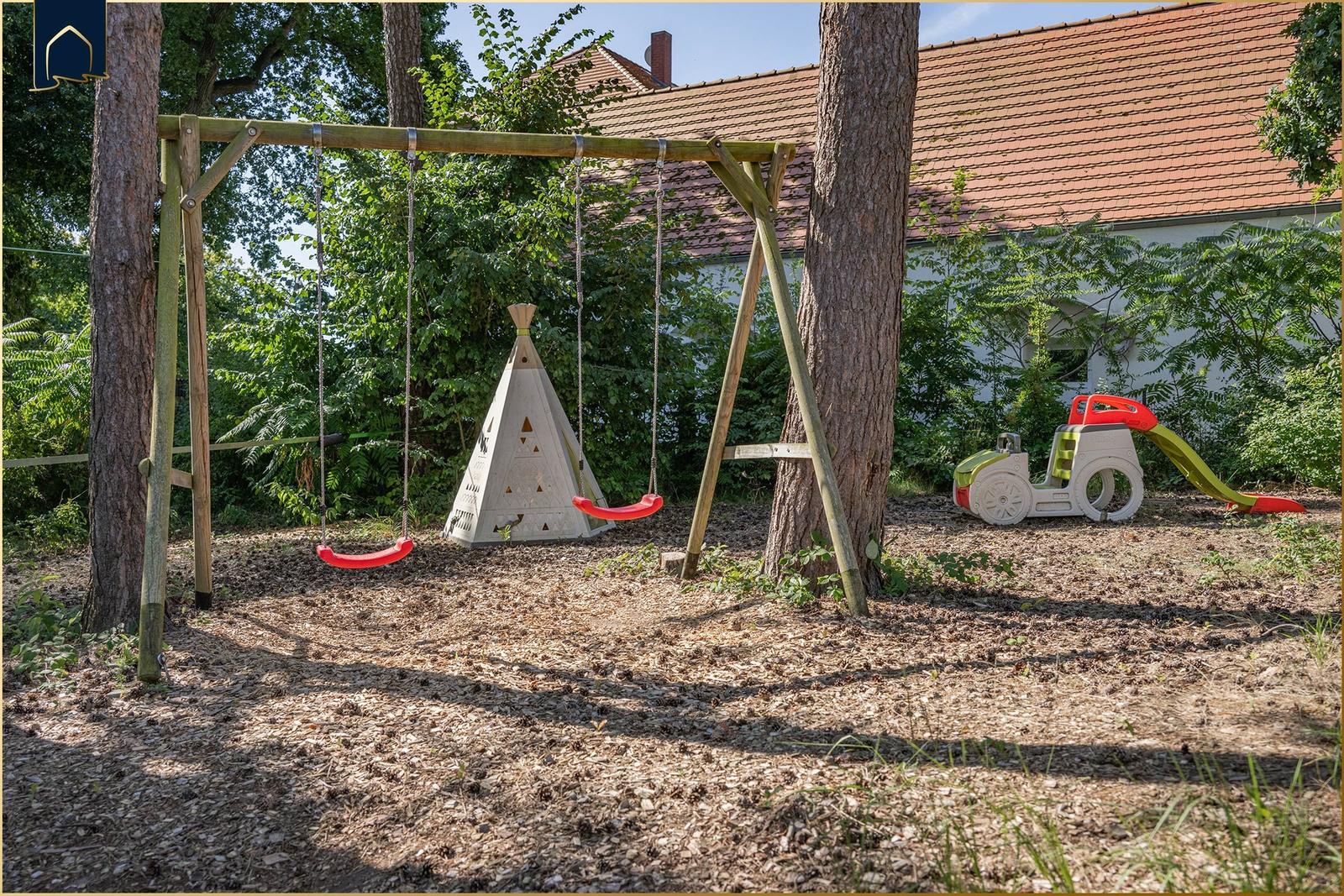 Holzüberdachter Bereich mit Grill, Tisch und Bänken. Hintergrund: Spielplatz und Bäume.