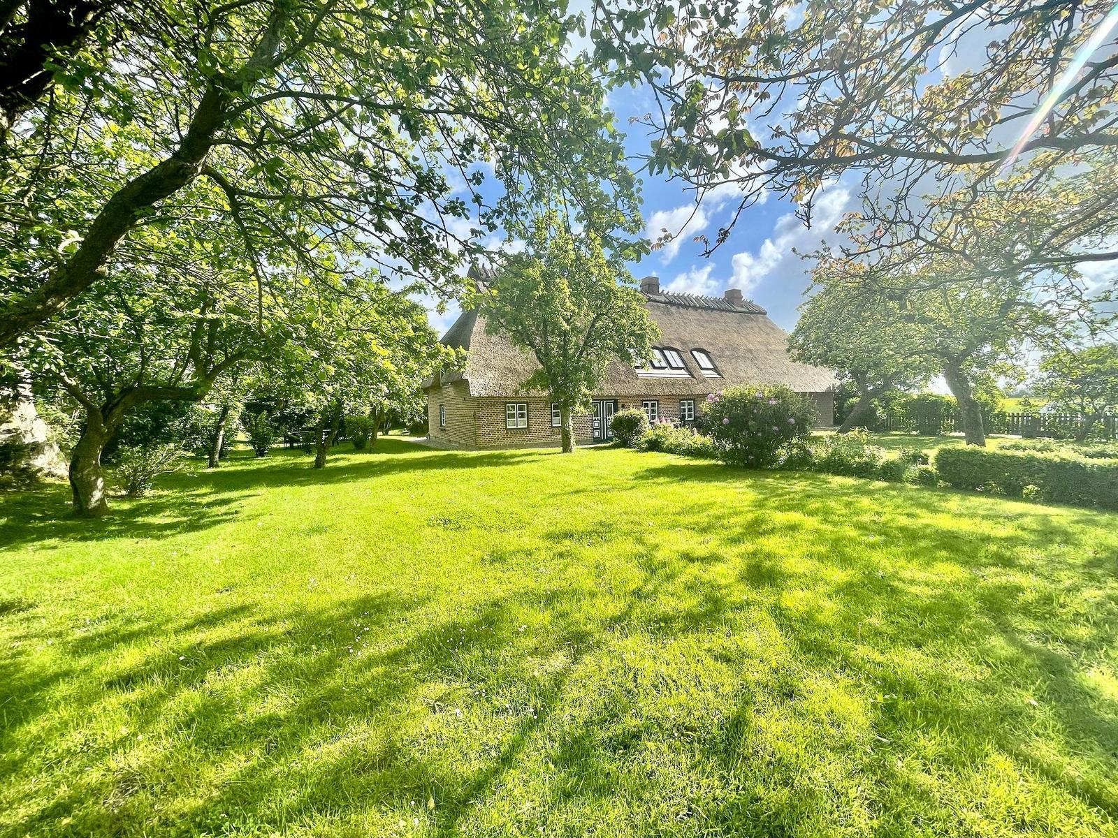 Haus mit Strohdach, grüner Rasen, Holzliege und Tisch mit Stühlen unter Bäumen.
