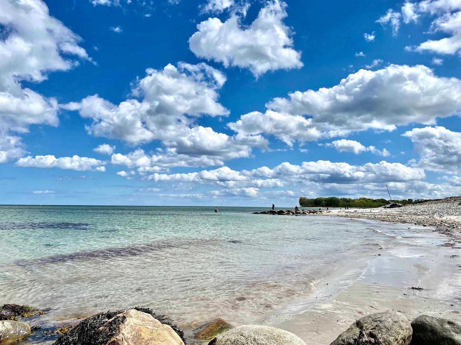 Strand mit flachem Wasser und Wellen, Horizont mit Bäumen und Häusern im Hintergrund.