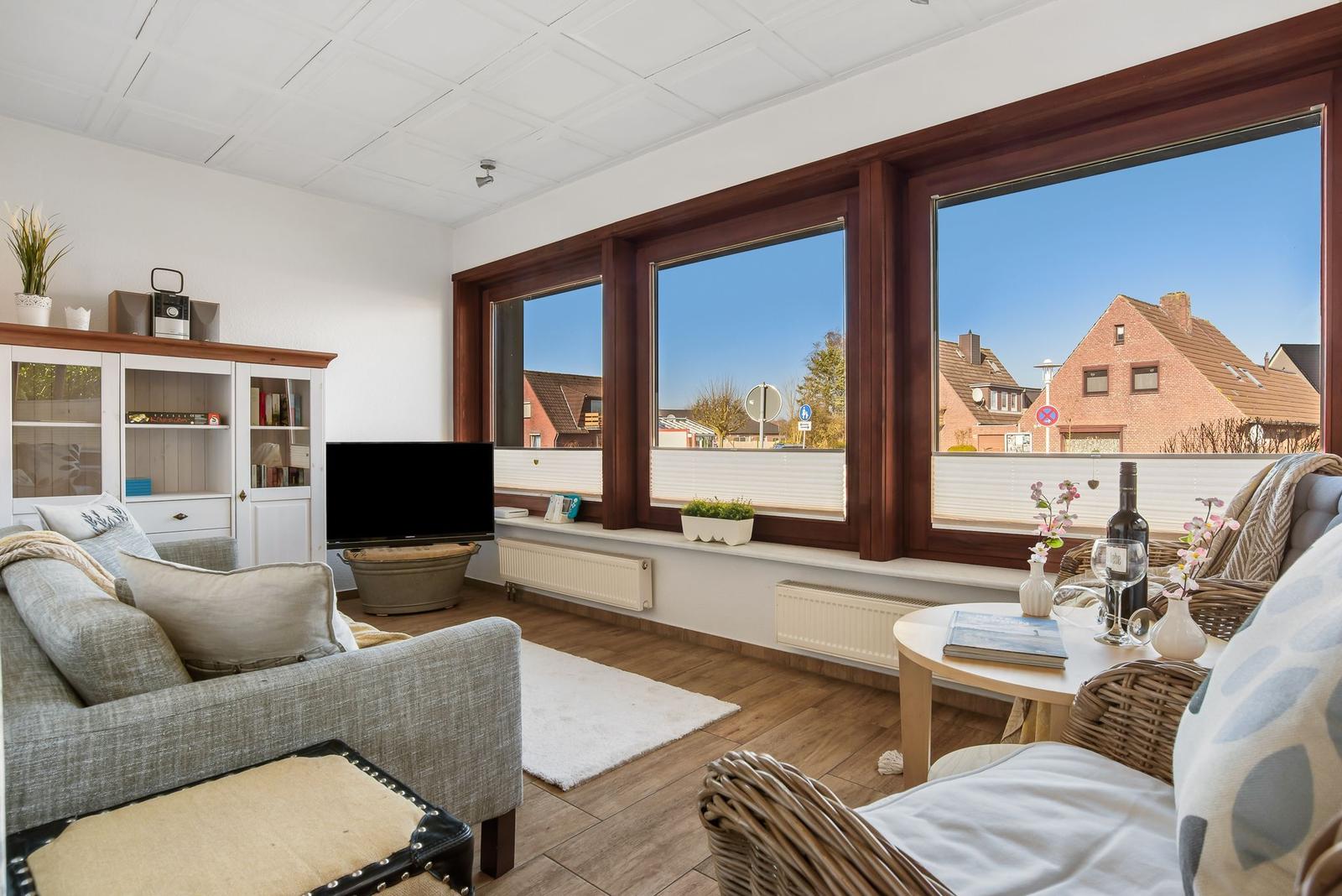 Wohnzimmer mit Sofa, Fernseher und Bücherregal. Große Fenster mit Blick nach draußen.