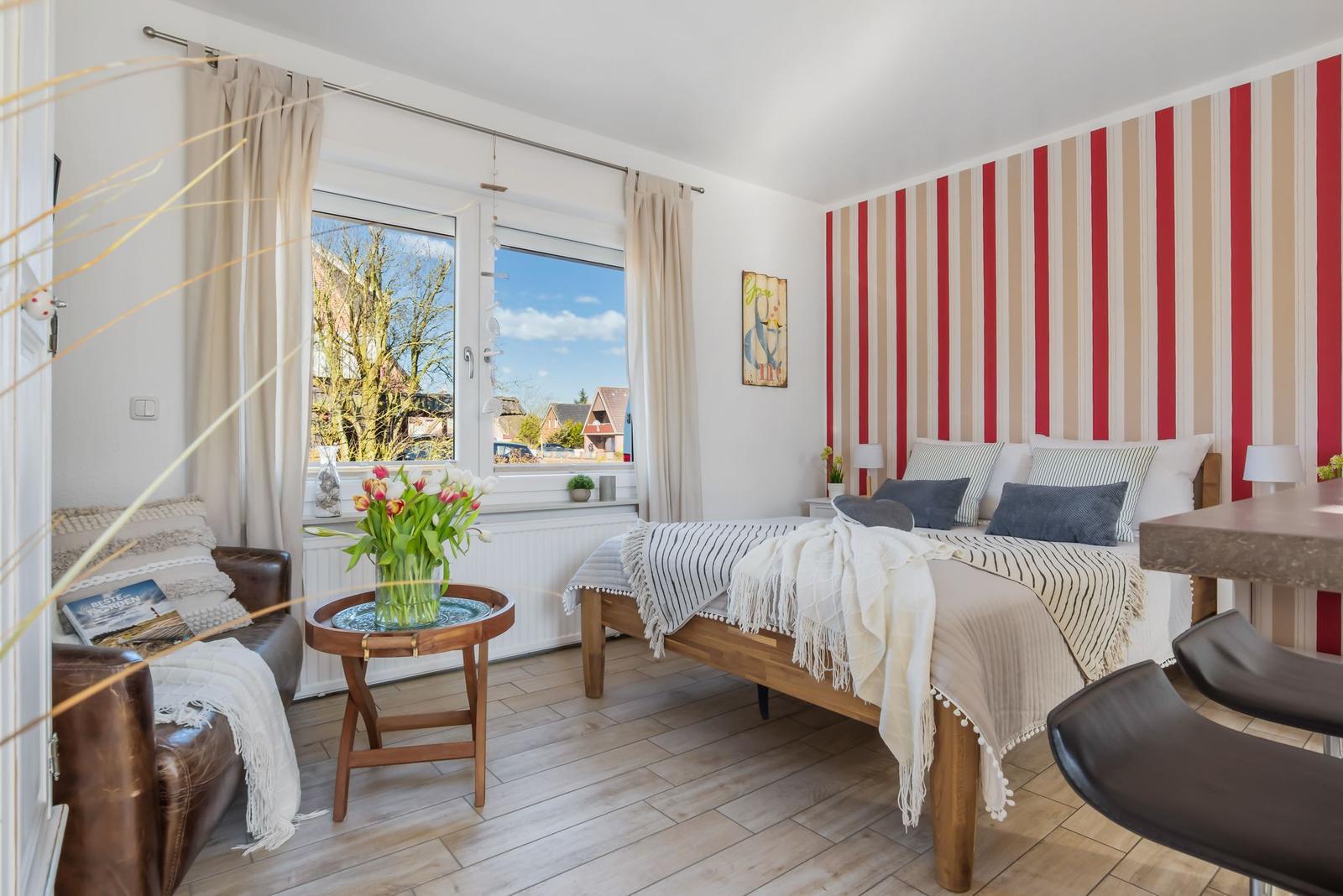 Schlafzimmer mit Streifenwand, Bett und Blick in die Küche mit Barhocker.