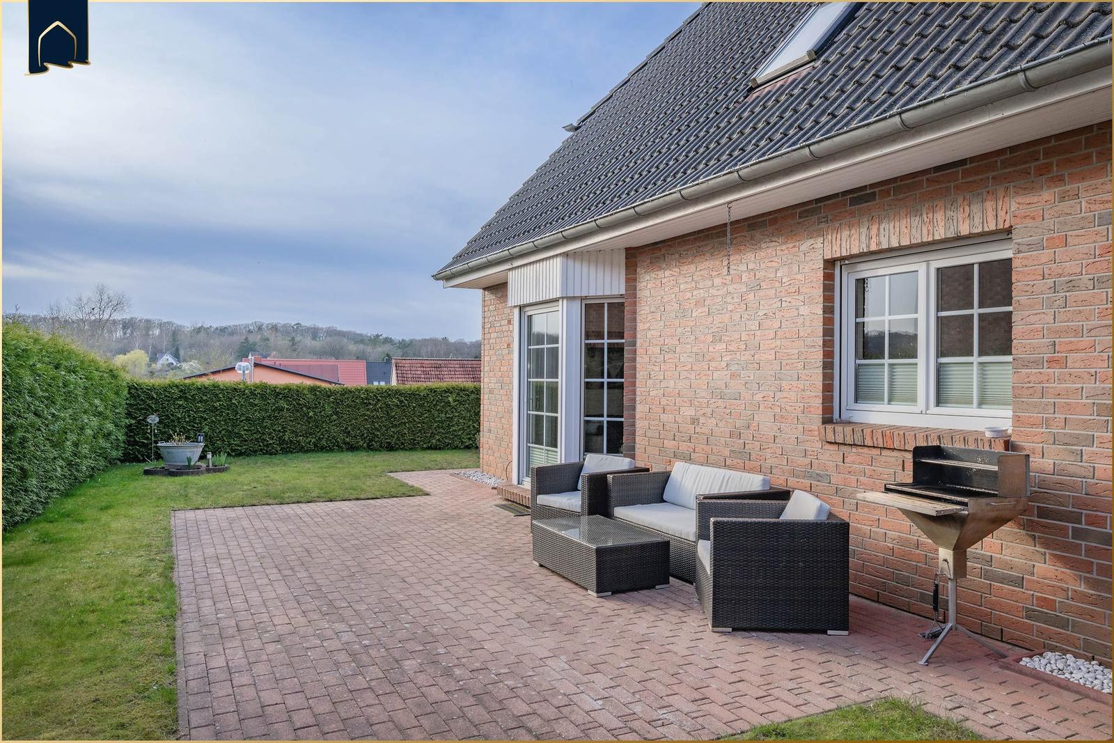 Ein Haus mit rotem Backstein und dunklem Dach, umgeben von grünen Hecken und einem gepflasterten Weg.