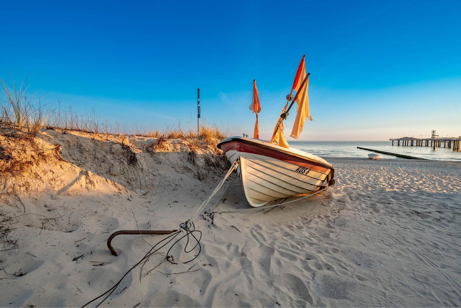 Eine weiße Bootsruhe mit KOS 1 liegt am Strand. Im Hintergrund erstreckt sich ein Steg ins Meer.