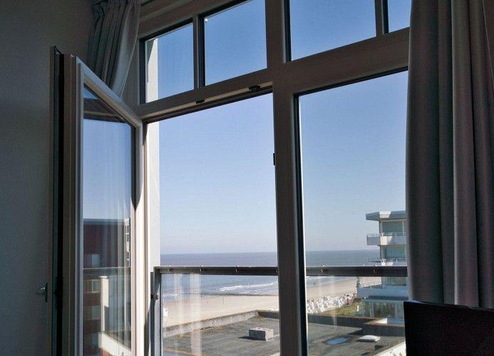 Durch die Glastüren führt ein Balkon mit Tisch. Im Hintergrund ist ein Haus mit rotem Dach sichtbar.