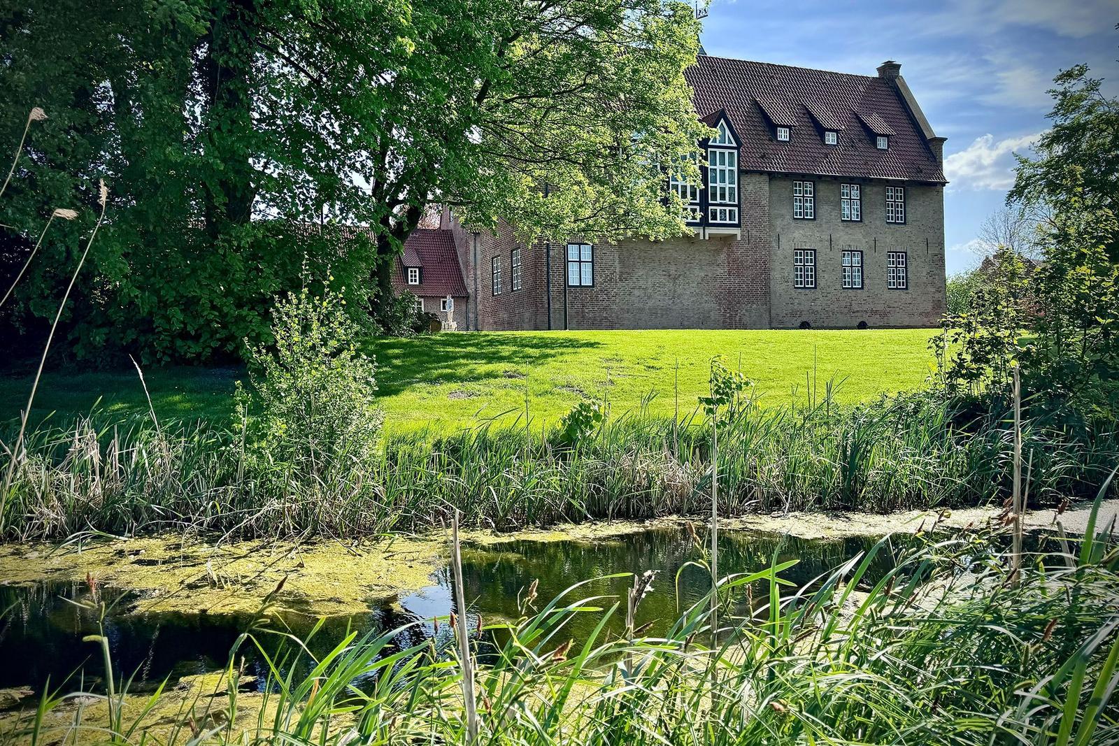 A canal with tall grass and trees, a stone bridge crosses the waterway.