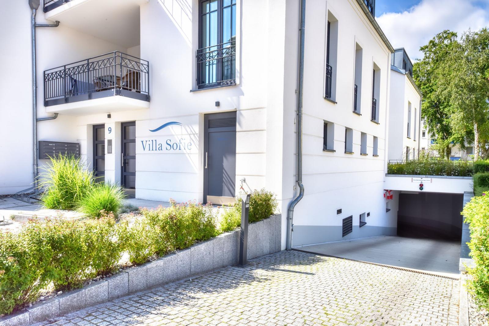 Weiße Villa mit Balkonen und begrüntem Vorgarten. Tiefgarage und Eingangsbereich sichtbar.