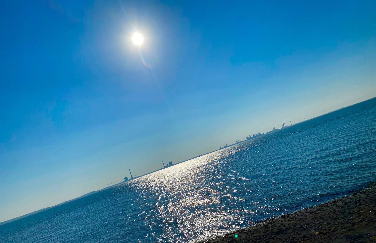 Steinpfad führt durch Wasser zu Horizont mit Schiffen und Sonne.