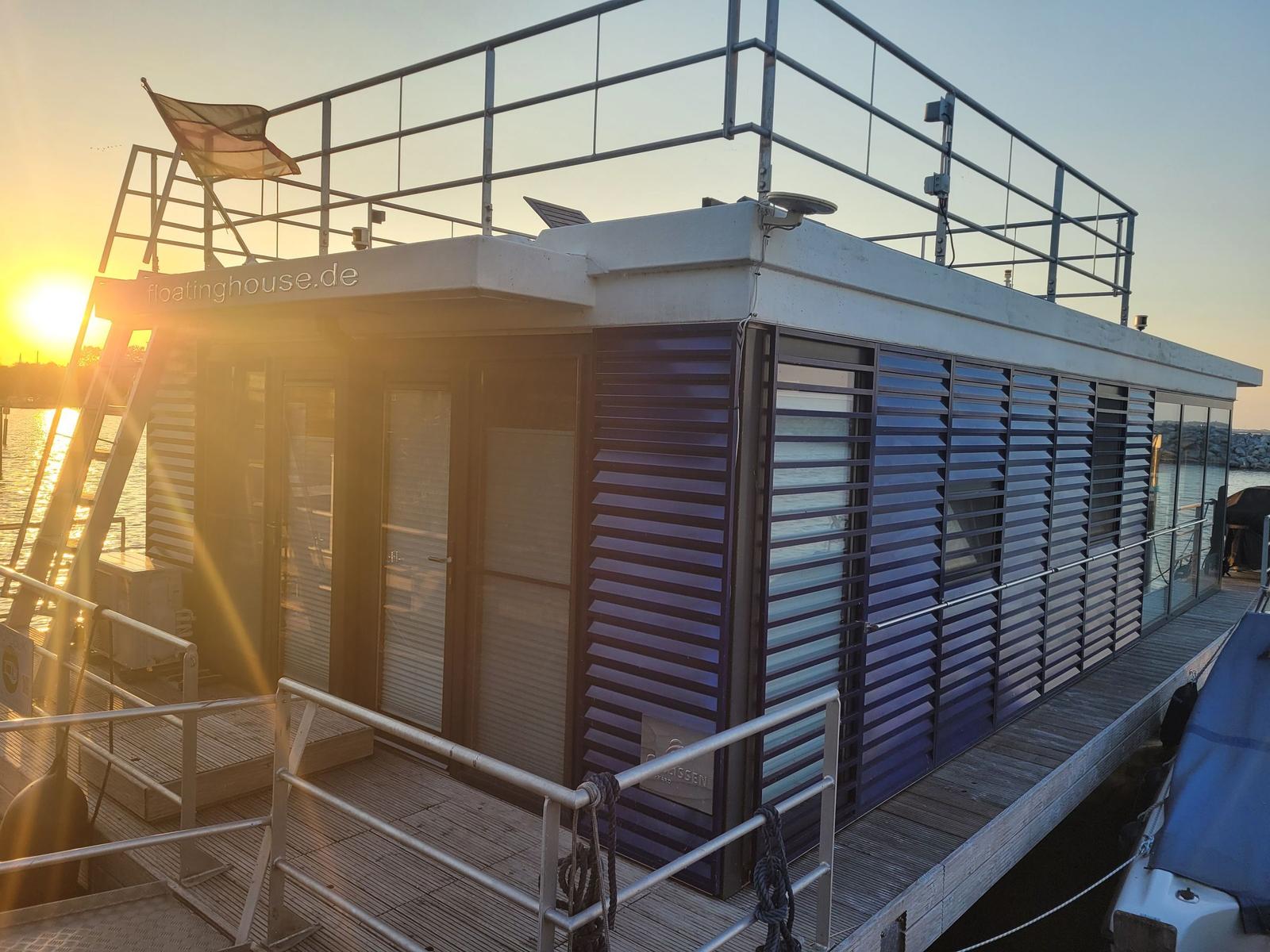 Holzsteg führt zu einem Hausboot am Wasser. Sonnenuntergang im Hintergrund.