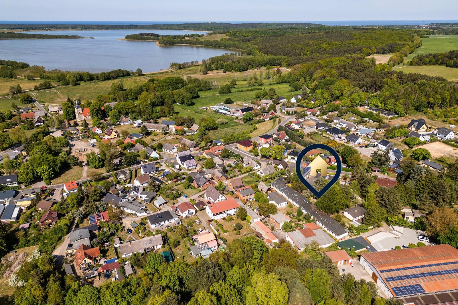Luftbild eines Dorfes mit Wäldern, Feldern und einem See im Hintergrund.