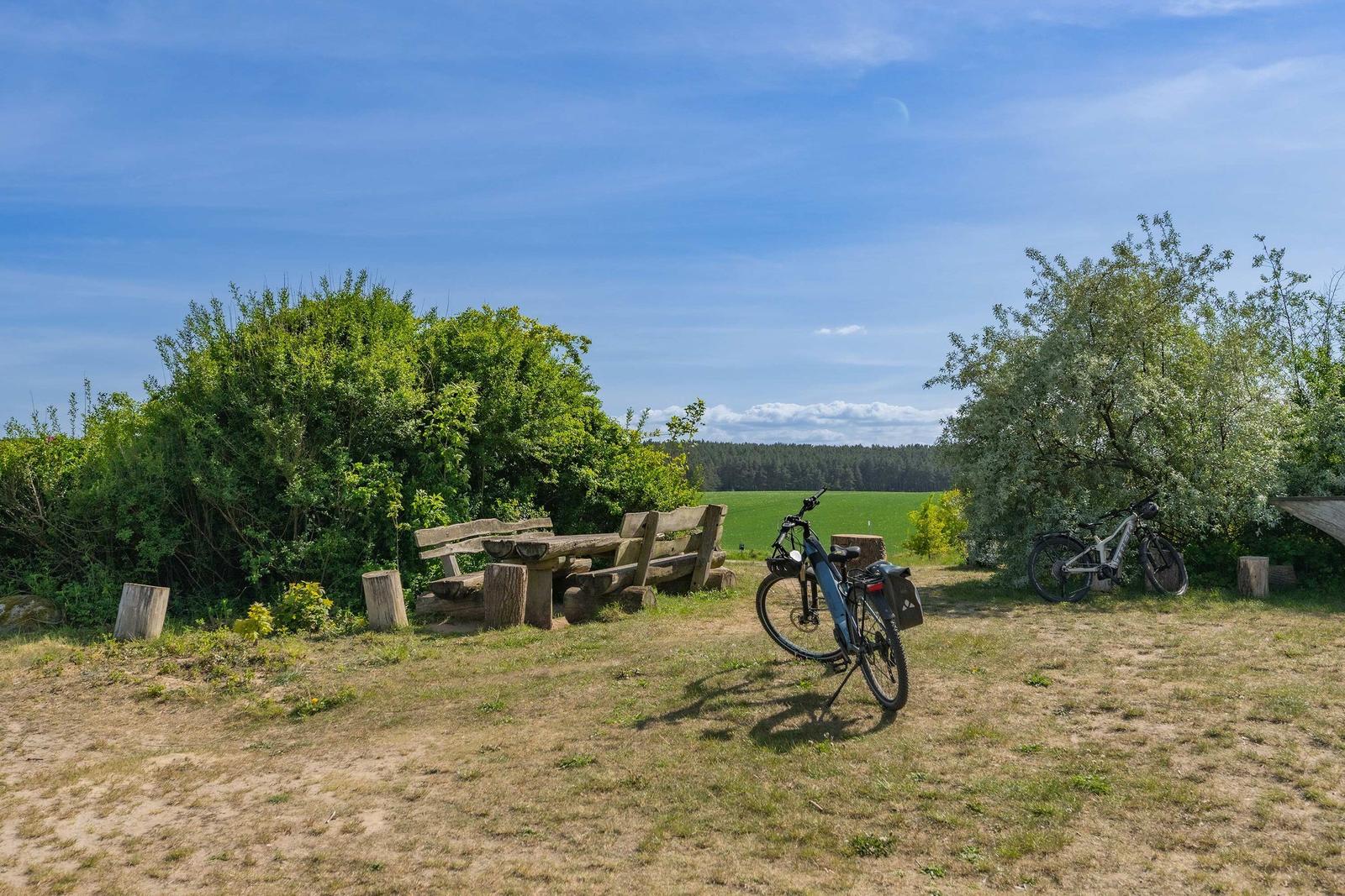 Holzmühle mit Sitzgruppe auf grünem Grund. Himmel blau, Bäume im Hintergrund.