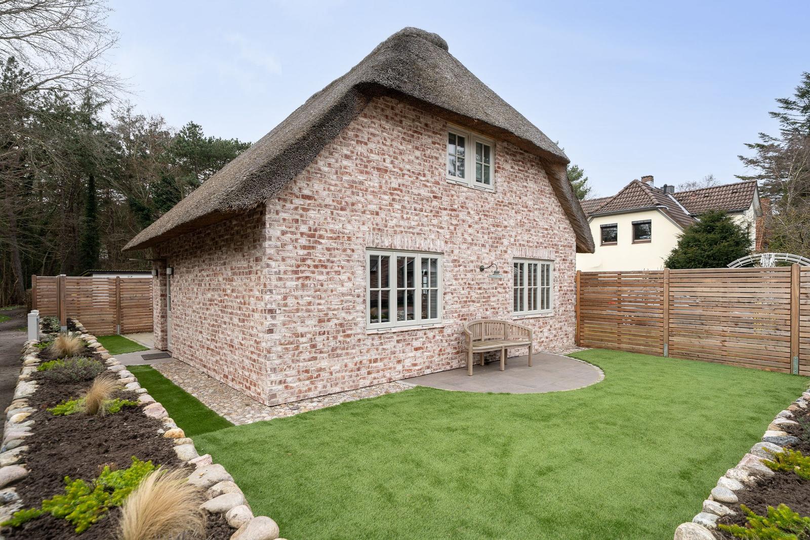 Ein Haus mit Strohdach und Steinmauer im Garten. Holzbank und Fenster sind sichtbar.