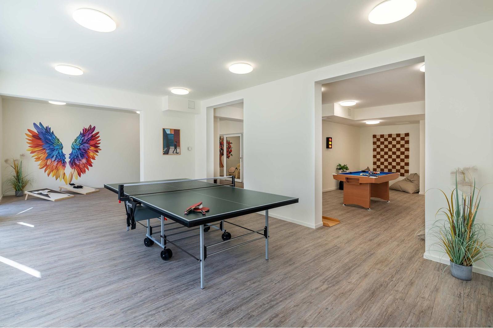 Raum mit Billardtisch, Sitzkissen und Pflanzen. Im Hintergrund ist ein Tischtennis-Tisch sichtbar.