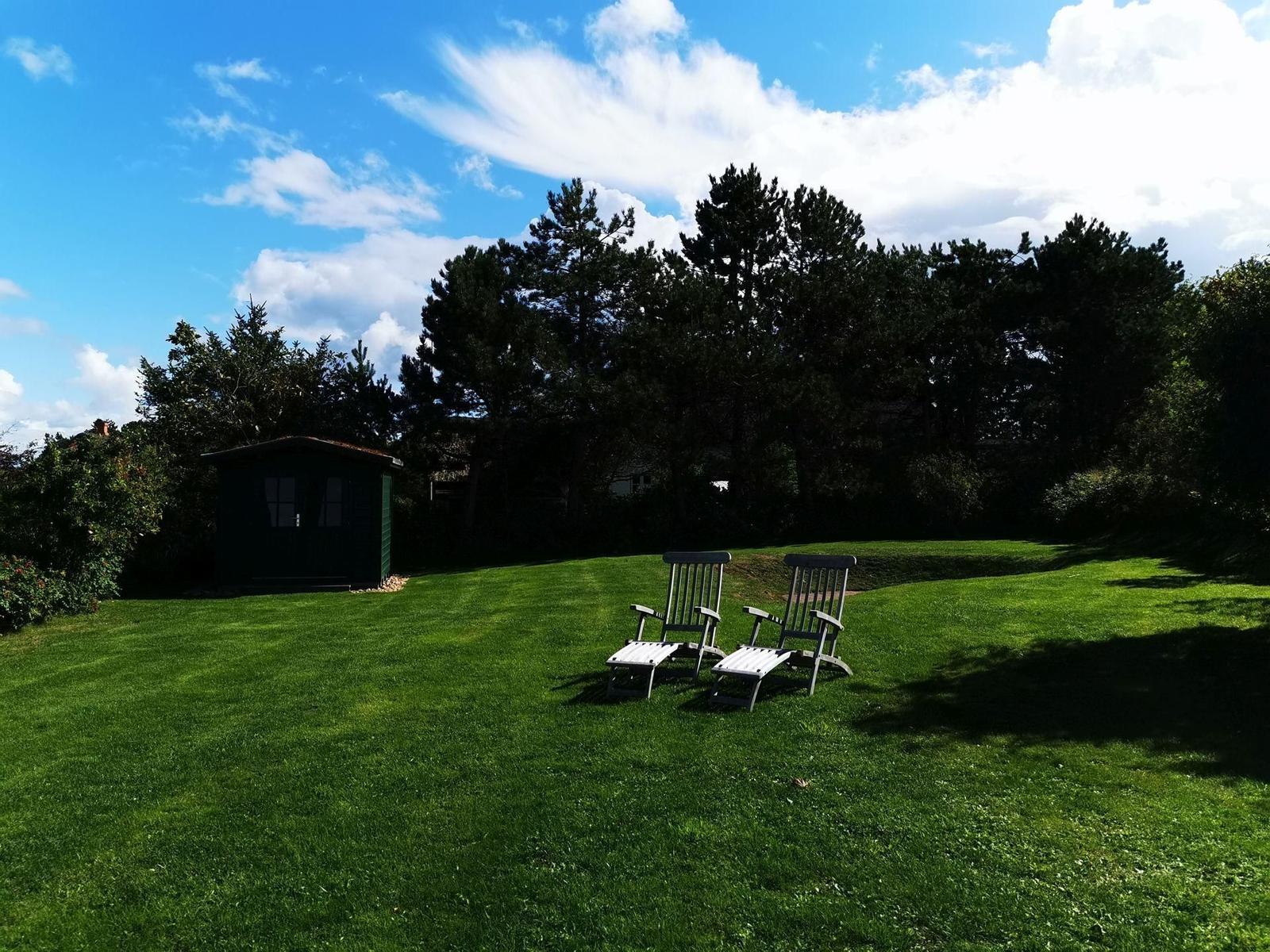 Garten mit Strandkorb, Holzstuhl und grünem Holzgebäude unter blauem Himmel.