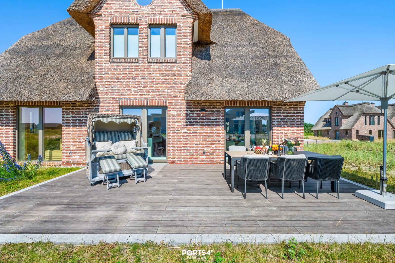 Terrasse mit Tisch, Stühlen und Sonnenschirm. Blick auf Strand und Häuser.