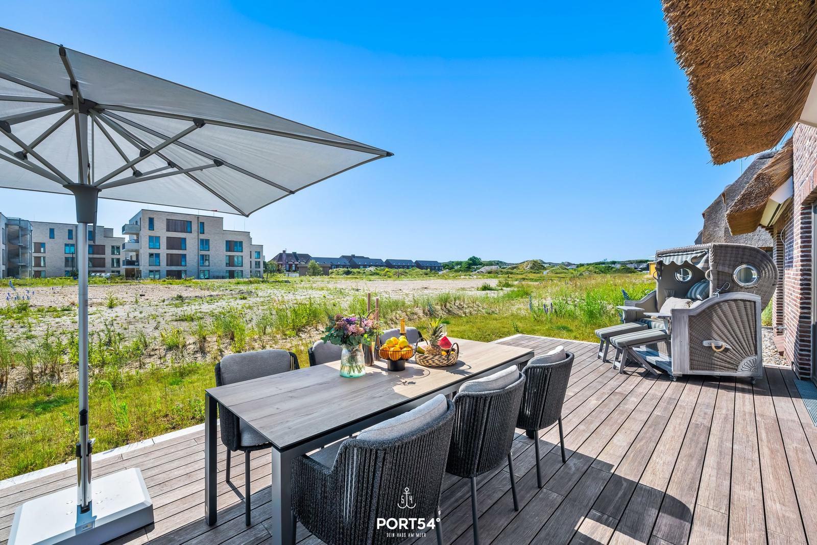 Terrasse mit Esstisch, Sonnenschutz und Strandkorb vor einem Haus mit Strohdach.