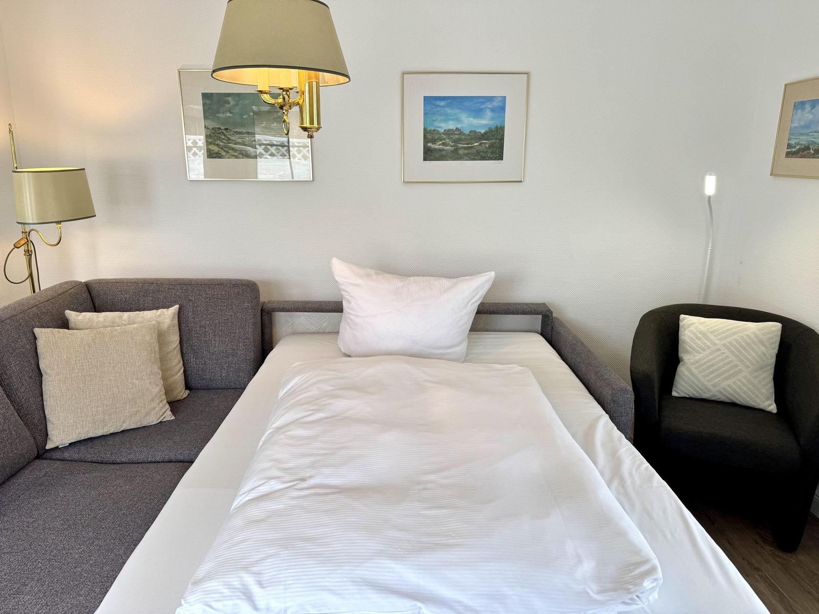 Grey sofa bed with white bedding, dark armchair and framed wall art.