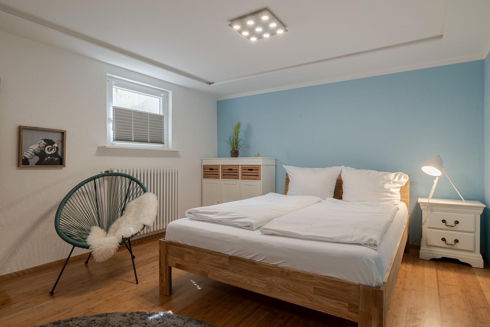 Schlafzimmer mit Holz-Doppelbett, hellblauer Wand und türkisem Sessel.